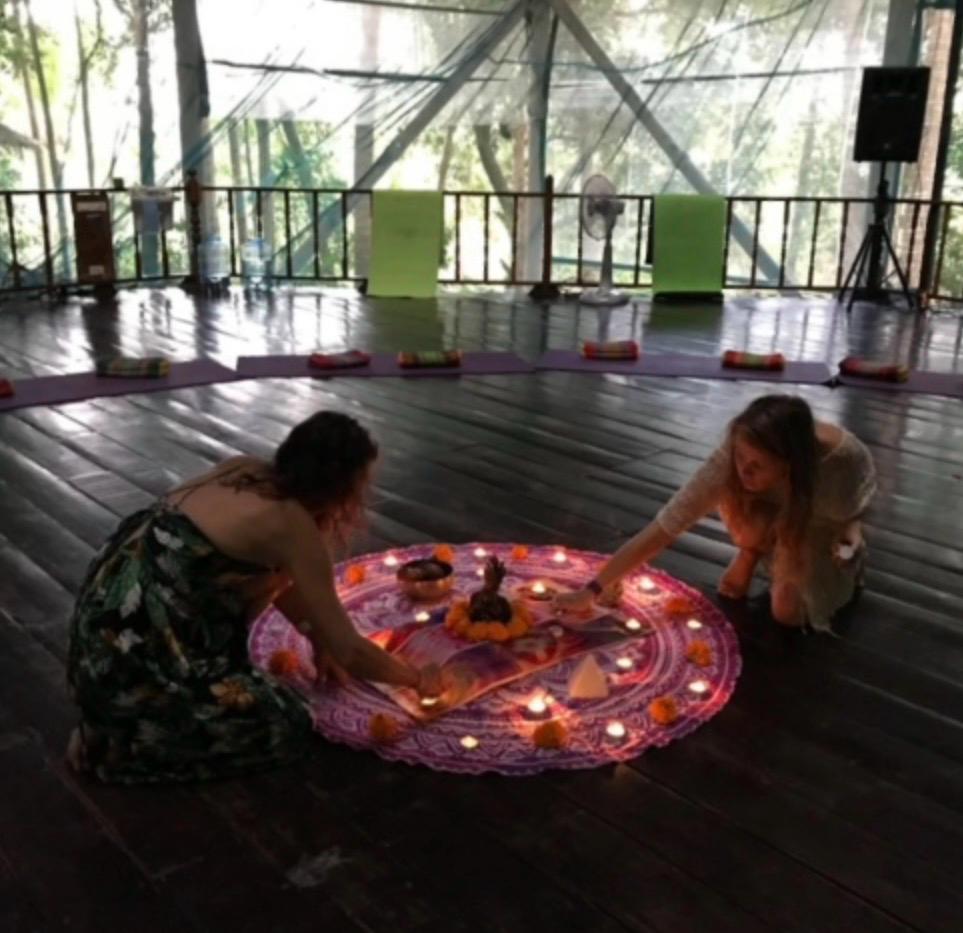 Due donne preparano un altare cerimoniale con mandala di candele e fiori al centro di una sala di legno