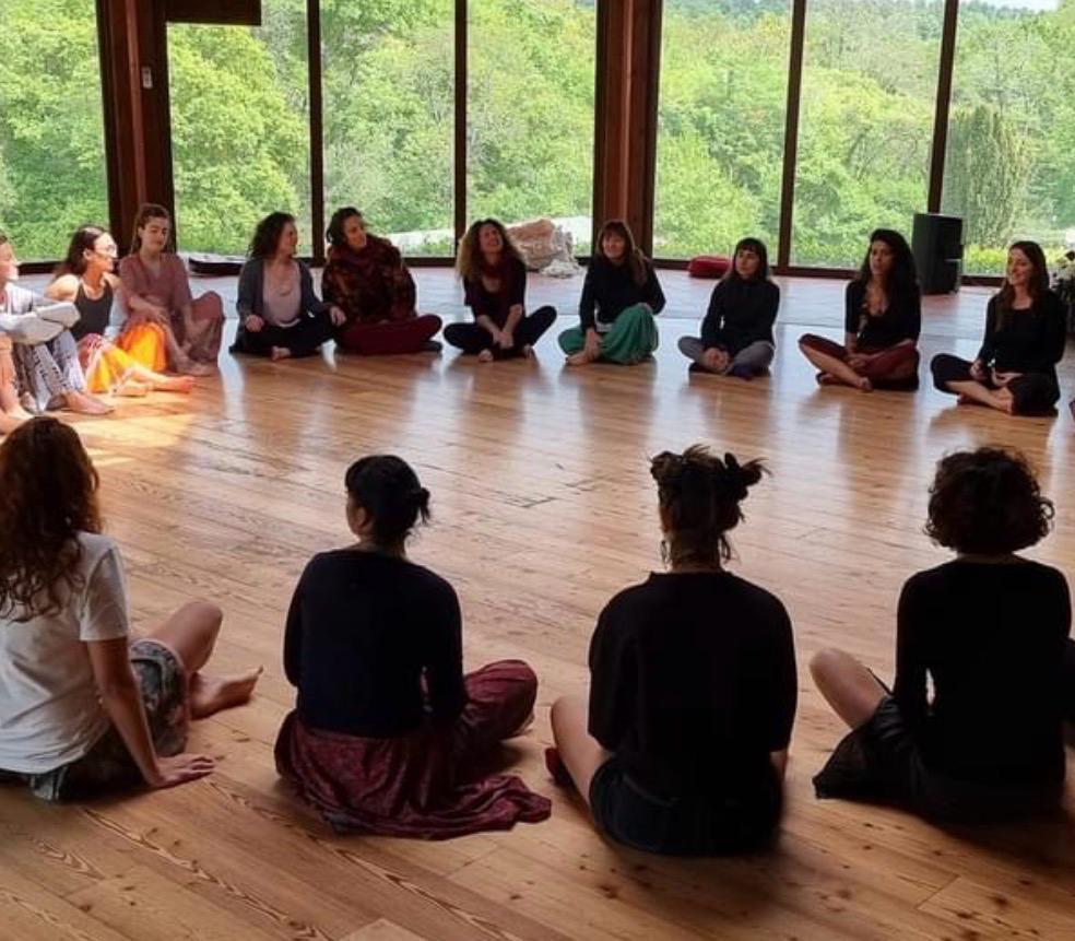 Cerchio di donne sedute a gambe incrociate in una sala con vetrate aperte sulla natura, pratica di gruppo meditativa