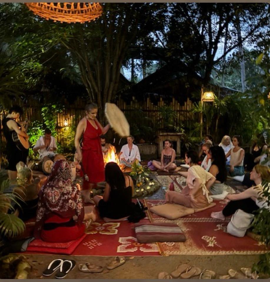 Cerimonia serale ancestrale all'aperto, donna in abito rosso suona un tamburo davanti al fuoco mentre un cerchio di partecipanti assiste